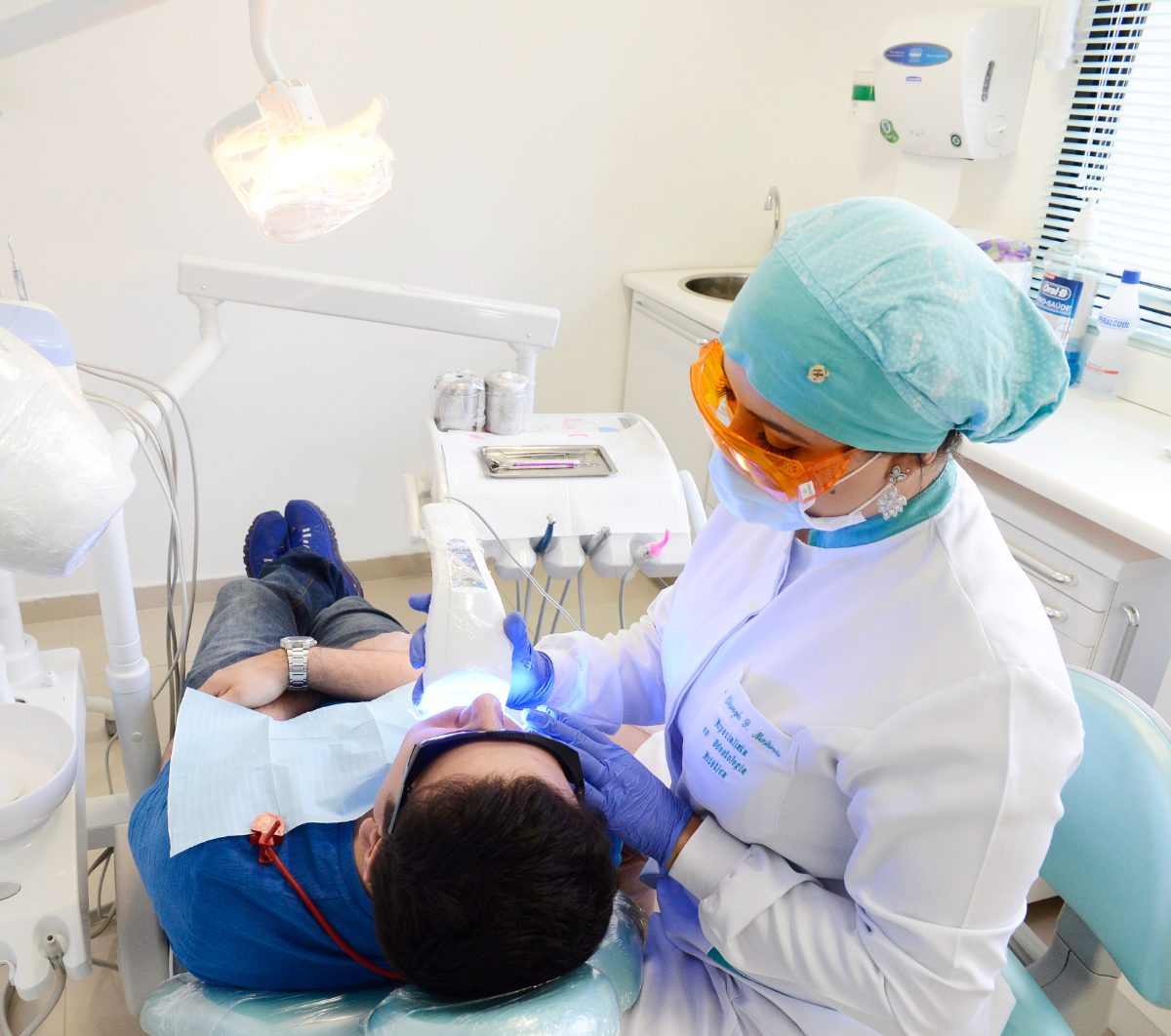 Dra. Elisangela atendendo um paciente na cadeira do consultório, com touca, óculos de proteção e luvas