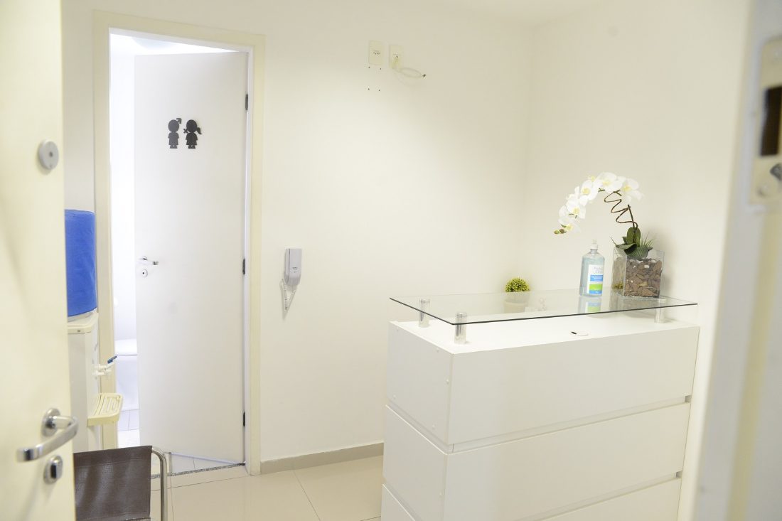 Recepção do consultório, branca, com balcão de vidro e orquídea